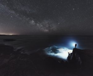 A man is silhouetted against the light of a bright spotlight as he searches the Atlantic under the Summer Milky Way. Long exposure.